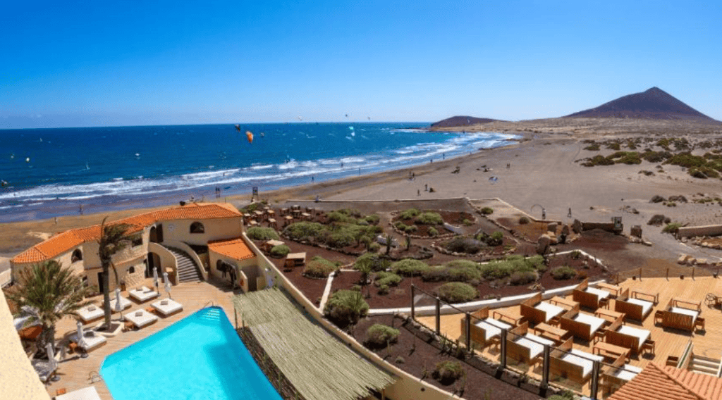 Hotel Playa Surf in El Médano, Tenerife. View from above looking at a pool with sun lounge chairs beyond which is ocean and kitesurfers.