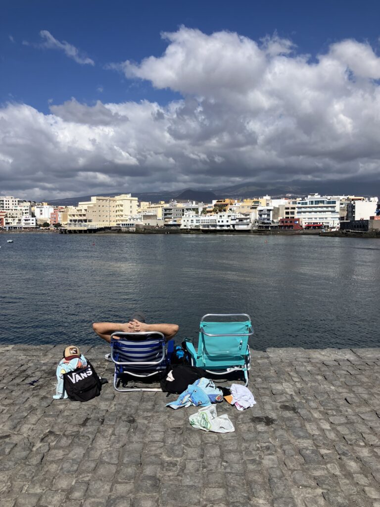 Man sitting in a low beach chair on a stone path, looking at the coastal town of El Médano