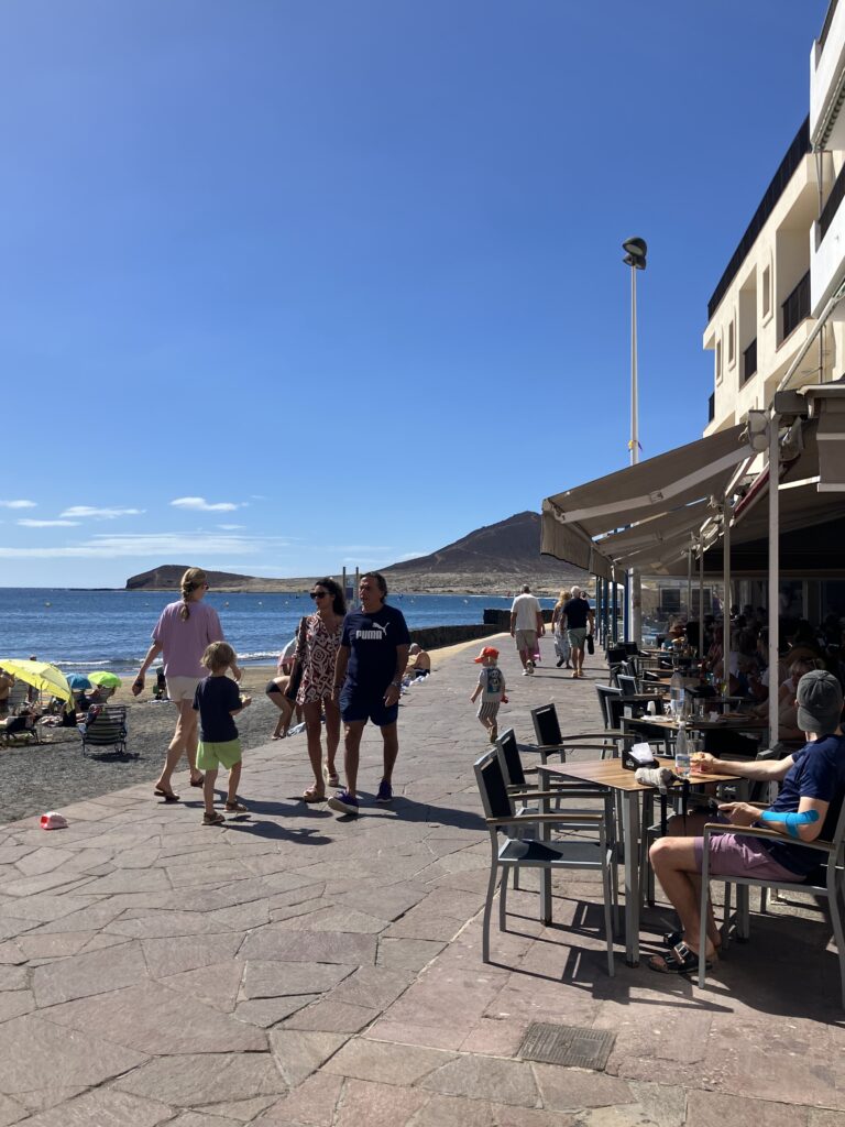 People walking along a stone seaside boardwalk. Man seated at a cafe overlooking the ocean.